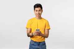 得快乐的年轻的亚洲纹身的家伙黄色的t恤持有智能手机微笑相机消息传递应用程序编辑照片帖子社会媒体