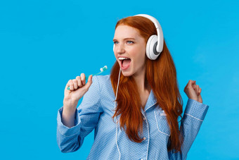 时髦的好看的红色头发的人女睡衣有趣的听音乐大白色耳机唱歌牙刷刷牙牙齿准备好了站蓝色的背景