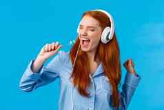 唱无忧无虑的兴奋快乐的红色头发的人女睡衣穿大耳机唱歌牙刷关闭眼睛享受太棒了音乐站蓝色的背景