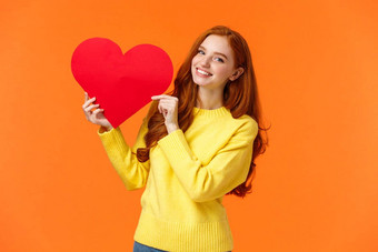 情人节一天完美的时间承认可爱的浪漫的温柔的红色头发的人女人黄色的毛衣持有大心标志微笑表达感情浪漫站橙色背景