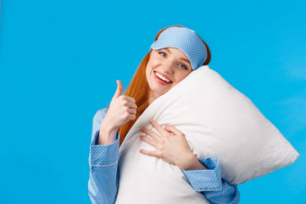 好健康的睡眠快乐的漂亮的红色头发的人女人睡眠面具睡衣拥抱软枕头显示竖起大拇指批准手势站很高兴蓝色的背景