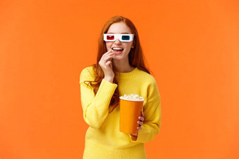 感兴趣逗乐微笑极客红色头发的人女人黄色的毛衣看电影电影穿眼镜吃爆米花站在好奇的娱乐兴奋表达式