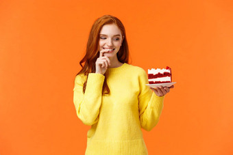 快乐的满意快乐红色头发的人女人撒谎日吃美味的食物持有美味的一块蛋糕咬唇微笑<strong>抵制</strong>诱惑欲望咬橙色背景