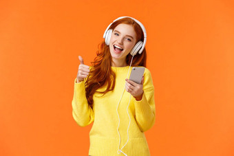 有吸引力的红色头发的人女孩收到圣诞节礼物很酷的耳机听音乐耳机显示翘拇指持有智能手机享受音乐唱歌微笑橙色背景