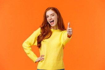 快乐的红色头发的人女人黄色的毛衣给翘拇指手势眨眼鼓励保持批准的想法表达积极的的意见推荐产品事件橙色背景