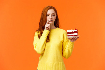 饮食健康的生活方式垃圾食物概念可爱的红色头发的人女人<strong>抵制</strong>诱惑咬美味的蛋糕持有甜点板咬唇思考卡路里橙色背景