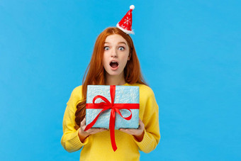 惊讶吓了一跳漂亮的红色头发的人女孩收到意想不到的礼物圣诞节同学开放口惊讶说不出话来盯着相机持有可爱的蓝色的现在包装盒子工作室背景