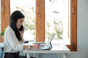 一边视图快乐亚洲女商人坐着工作场所办公室年轻的女人工作金融文档数据图表移动PC调用电话办公室