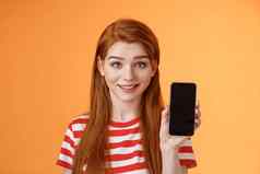 特写镜头可爱的高加索人红色头发的人女孩显示智能手机屏幕橙色背景微笑可爱的推荐应用程序促进应用程序在线提供出售介绍移动电话