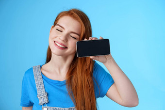 无忧无虑的温柔的红色头发的人欧洲女倾斜头微笑关闭眼睛持有智能手机水平显示爱的女朋友照片浪漫的视频在线站蓝色的背景快乐的