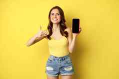 快乐微笑时尚的女孩显示拇指智能手机应用程序接口移动电话显示满意网站在线购物应用程序站黄色的背景