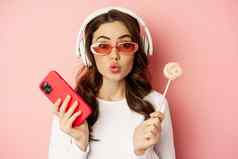 时尚的年轻的女人萝莉波普手机穿太阳镜耳机听音乐站粉红色的背景