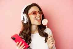 美丽的女模型听音乐耳机持有萝莉波普移动电话摆姿势太阳镜站粉红色的背景