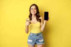 年轻的时尚的女人显示移动电话屏幕指出智能手机显示应用程序接口在线商店微笑很高兴站黄色的背景