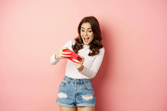 快乐美丽的女孩玩移动视频游戏持有智能手机水平看手机兴奋脸粉红色的背景