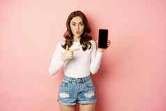 惊讶浅黑肤色的女人女孩指出移动电话应用程序屏幕显示智能手机广告在线购物提供站惊讶粉红色的背景