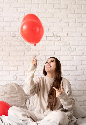 年轻的浅黑肤色的女人女人坐着床上庆祝情人节一天持有红色的心气球