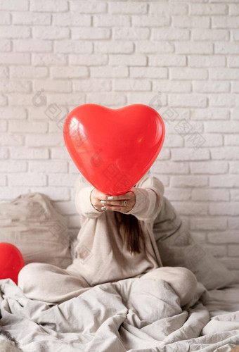 年轻的浅黑肤色的女人女人坐着床上庆祝情人节一天持有红色的心气球