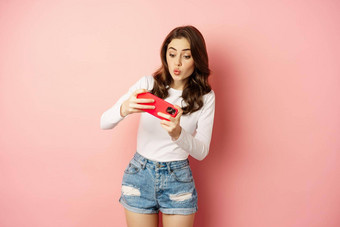 快乐美丽的女孩玩移动视频游戏持有智能手机水平看手机兴奋脸粉红色的背景