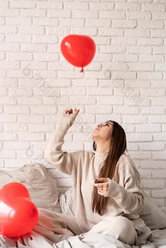 年轻的浅黑肤色的女人女人坐着床上庆祝<strong>情人节</strong>一天持有红色的心气球