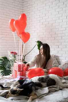 年轻的有趣的女人坐着床上庆祝情人节一天包装礼物