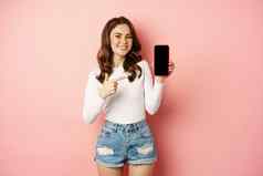 骄傲快乐微笑女孩指出手指显示移动电话屏幕手机应用程序促销文本智能手机站粉红色的背景