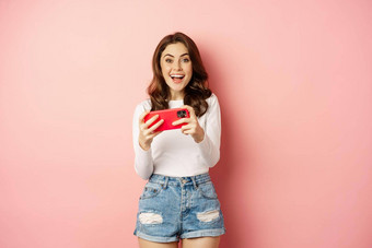 快乐美丽的女孩笑微笑看视频玩游戏移动电话水平智能手机站粉红色的背景