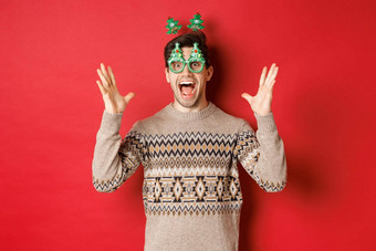 图像兴奋惊讶高加索人的家伙聚会，派对眼镜圣诞节毛衣提高手使大公告享受一年庆祝活动红色的背景