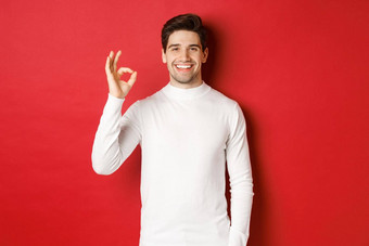 概念冬天假期圣诞节生活方式快乐英俊的男人。白色毛衣显示标志微笑很高兴推荐商店站红色的背景