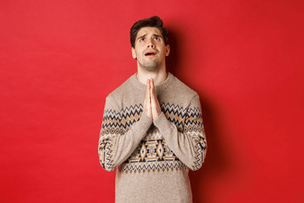 图像紧张充满希望的男人。祈祷神乞讨圣诞节穿冬天毛衣恳求站红色的背景