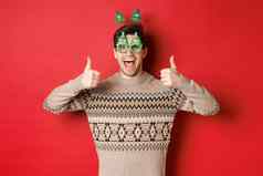 图像兴奋英俊的的家伙聚会，派对眼镜圣诞节毛衣显示竖起大拇指赞扬伟大的一年庆祝活动站红色的背景