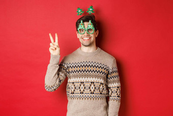 肖像有趣的可爱的男人。聚会，派对眼镜圣诞节毛衣微笑显示和平标志摆姿势红色的背景一年广告