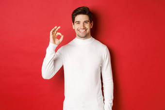 肖像快乐满意英俊的男人。穿白色毛衣显示标志微笑批准好推荐商店站红色的背景