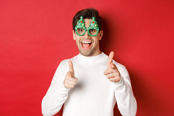 快乐的高加索人的家伙聚会，派对眼镜白色毛衣微笑指出手指相机祝快乐圣诞节快乐一年站红色的背景