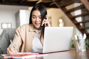 微笑亚洲女孩工作首页会说话的客户端回答电话调用采取笔记笔记本坐着移动PC