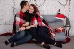 快乐夫妇拥抱灰色沙发上覆盖红色的网纹格子毯红色的圣诞节装饰舒适的年轻的家庭