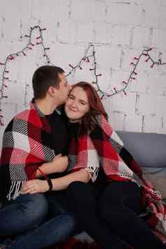 快乐夫妇拥抱灰色沙发上覆盖红色的网纹格子毯红色的圣诞节装饰舒适的年轻的家庭