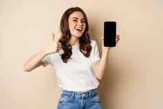 图像快乐年轻的女人显示拇指移动电话屏幕推荐网上商店在线购物应用程序站很高兴米色背景