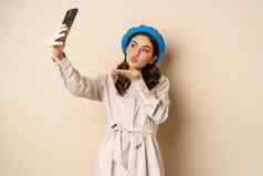 美丽的现代女人摆姿势自拍持有智能手机采取图片照片移动电话站快乐米色背景