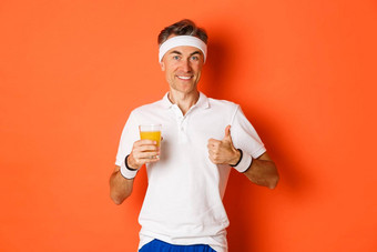 概念锻炼健身房生活方式图像适合中年男人。住形状练习健康的食物持有新鲜的汁显示竖起大拇指站橙色背景