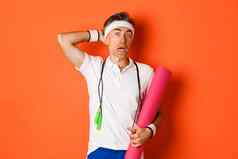 概念锻炼健身房生活方式图像尴尬的中年运动员困惑持有瑜伽席跳过绳子站橙色背景
