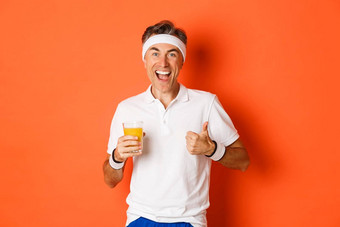 概念锻炼健身房生活方式图像活跃的健康的中年体育运动的家伙显示竖起大拇指喝汁微笑快乐站橙色背景