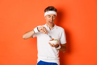 概念体育<strong>运动健身</strong>生活方式肖像快乐的中年的家伙锻炼统一的持有叉沙拉吃健康的食物站橙色背景