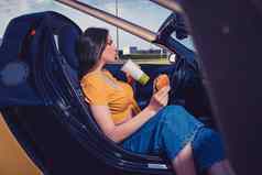 夫人蓝色的牛仔裤橙色前喝饮料纸杯持有汉堡坐着黄色的车Cabrio快食物模拟