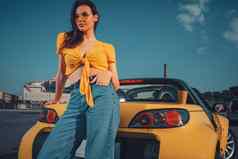 女孩蓝色的牛仔裤橙色前太阳镜摆姿势倾斜黄色的车蓬式汽车纸杯饮料树干停车很多复制空间