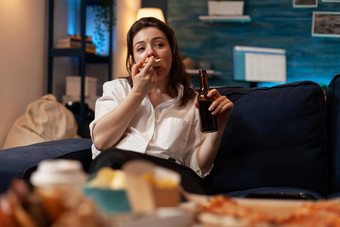 女人吃片热披萨交付坐着沙发上持有啤酒瓶电视