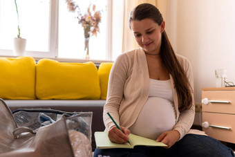 可爱的怀孕了女人写作包装列表孕妇医院笔记本准备袋年轻的莱迪怀孕有趣的花时间地板上沙发<strong>首页母亲</strong>医学健康护理概念
