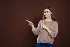 微笑快乐的女人宽松的毛衣摆姿势棕色（的）工作室墙指出复制空间广告促销文本积极的情绪感情快乐幸福