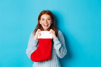 冬天假期礼物概念快乐十几岁的红色头发的人女孩接收圣诞节礼物开放圣诞节长袜微笑惊讶站蓝色的背景