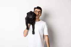 快乐的赶时髦的人的家伙眼镜t恤携带可爱的黑色的哈巴狗肩膀微笑狗老板玩宠物站白色背景